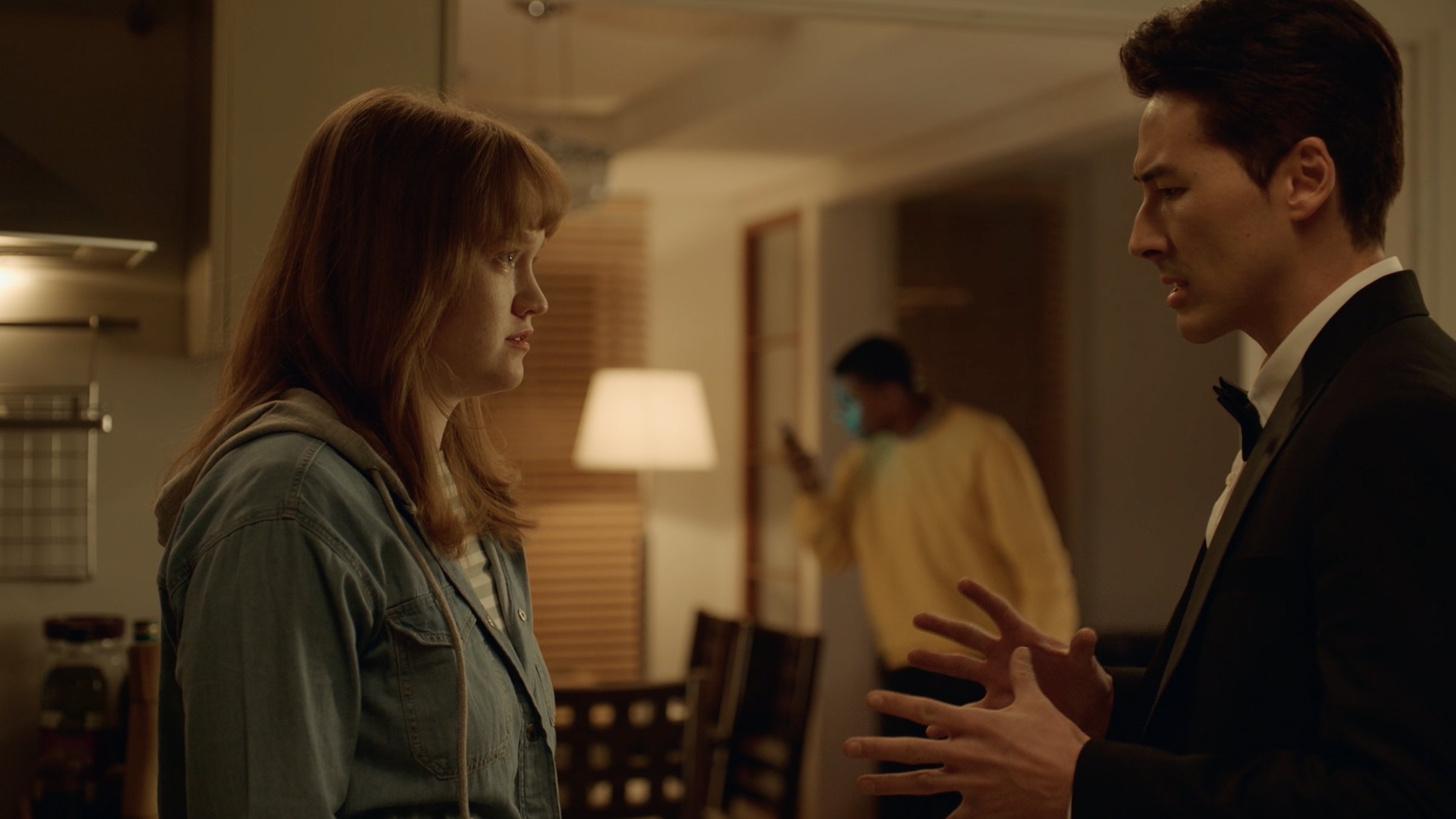 Claire and Joon face off in a kitchen while a man in a yellow sweater lurks in the background