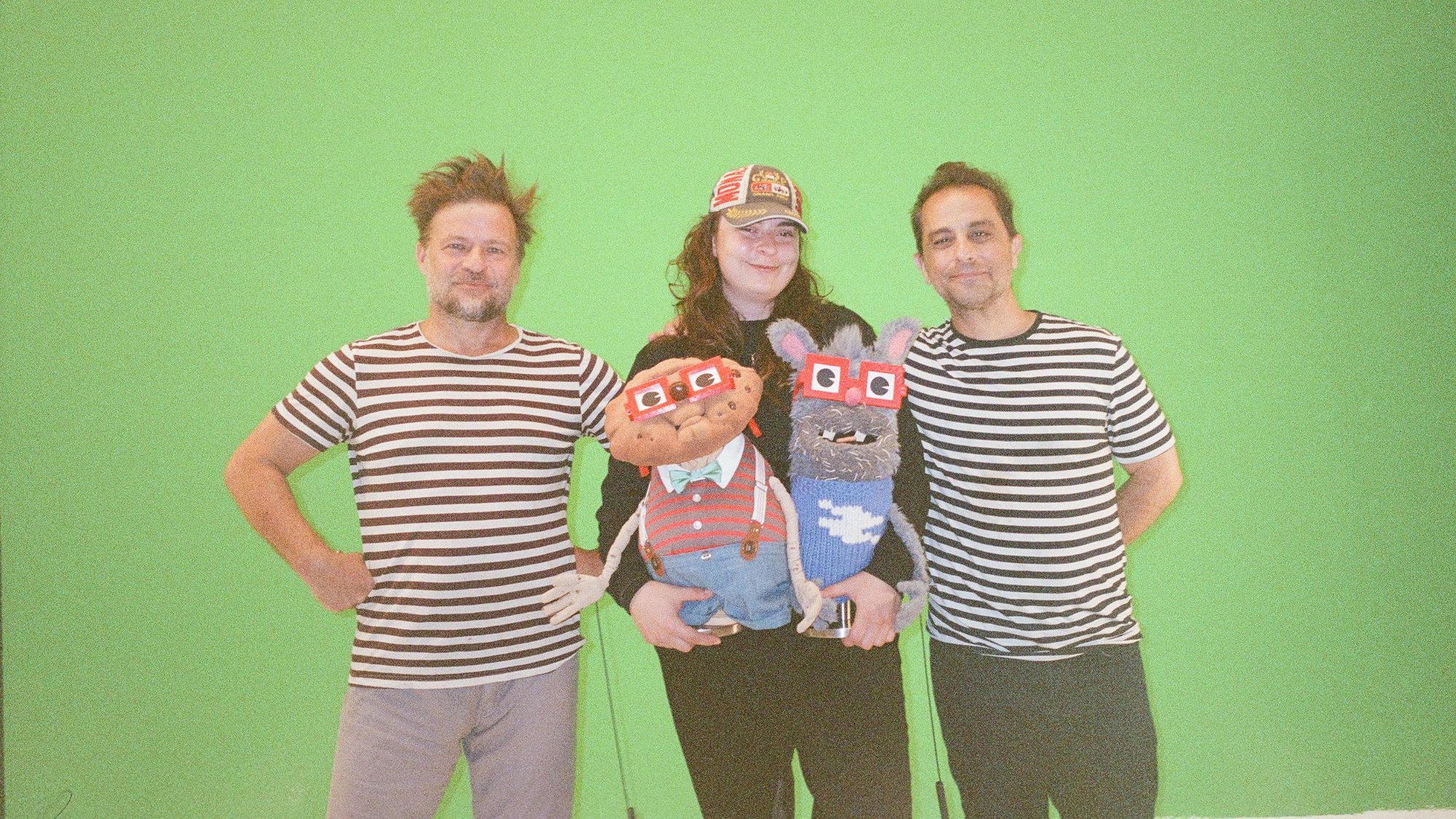 Film photo on the green-screen stage: three members of the team, two in matching striped shirts, posing with Chip and Benny